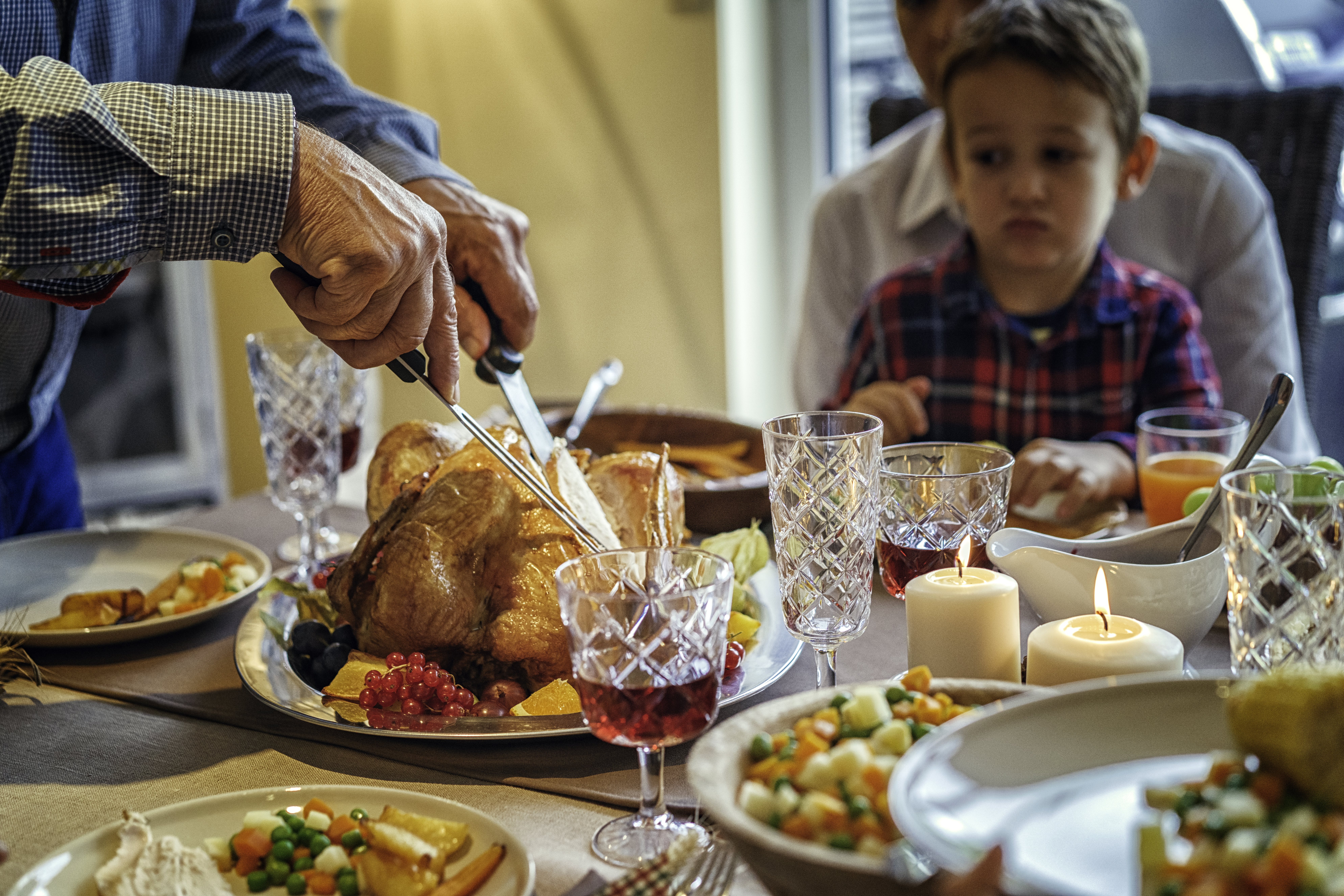 Grandfather carving stuffed turkey for holiday dinner with his family. The Thanksgiving Dinner is served with roasted potatoes, cranberry sauce, vegetables and red berry jelly.
